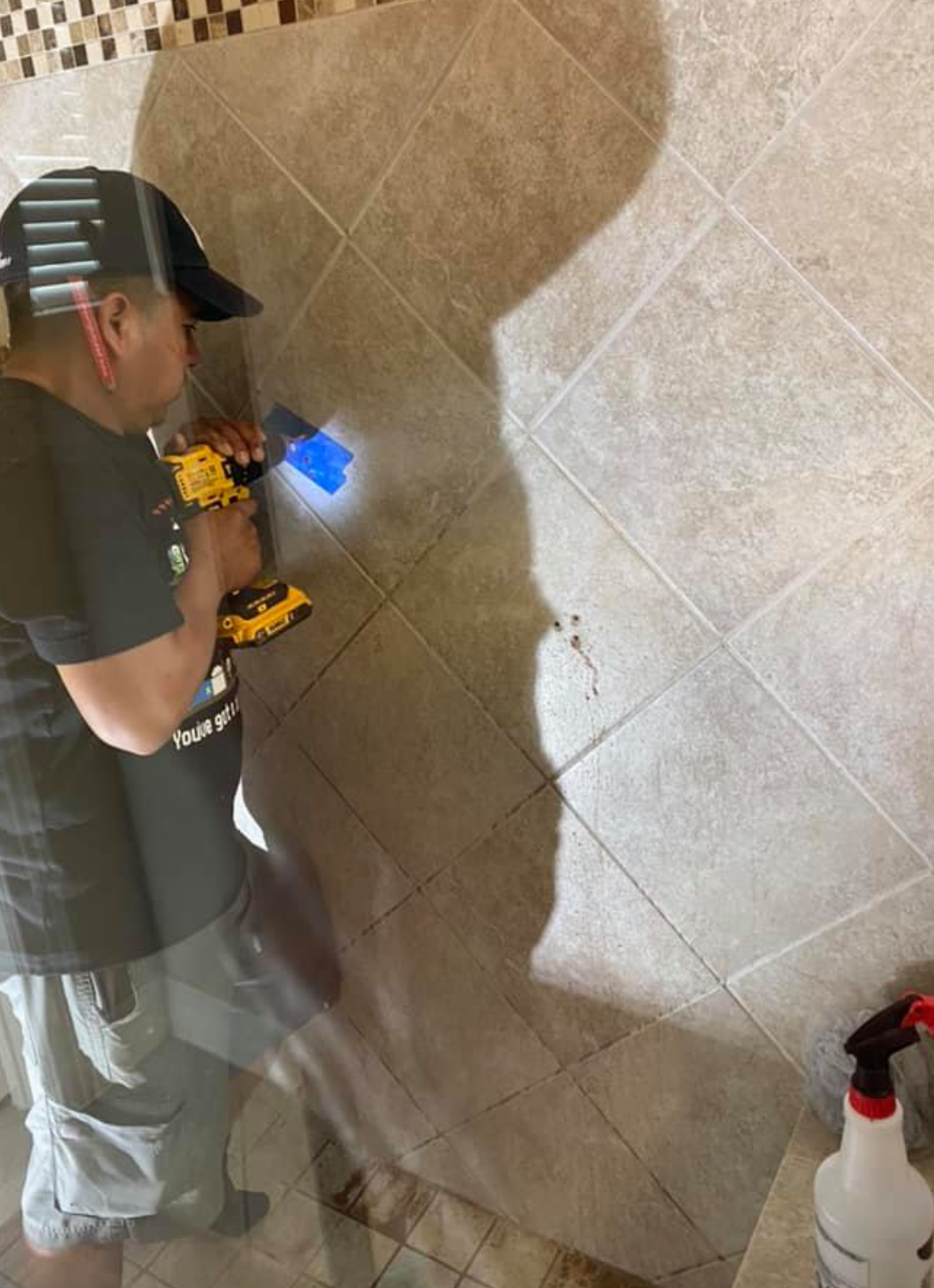 Man drilling into tile in a shower. He wears a cap, black shirt, gray pants. Bright light from the drill.
