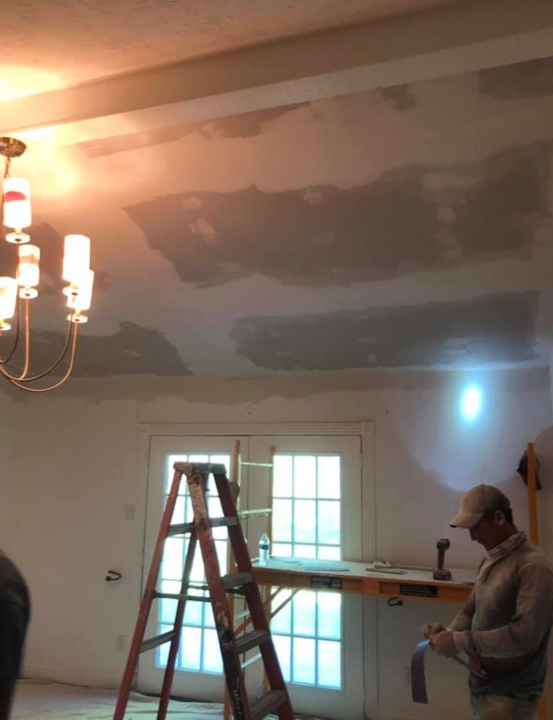 Man repairs ceiling with patches; ladder in front of french doors, chandelier, bright light.