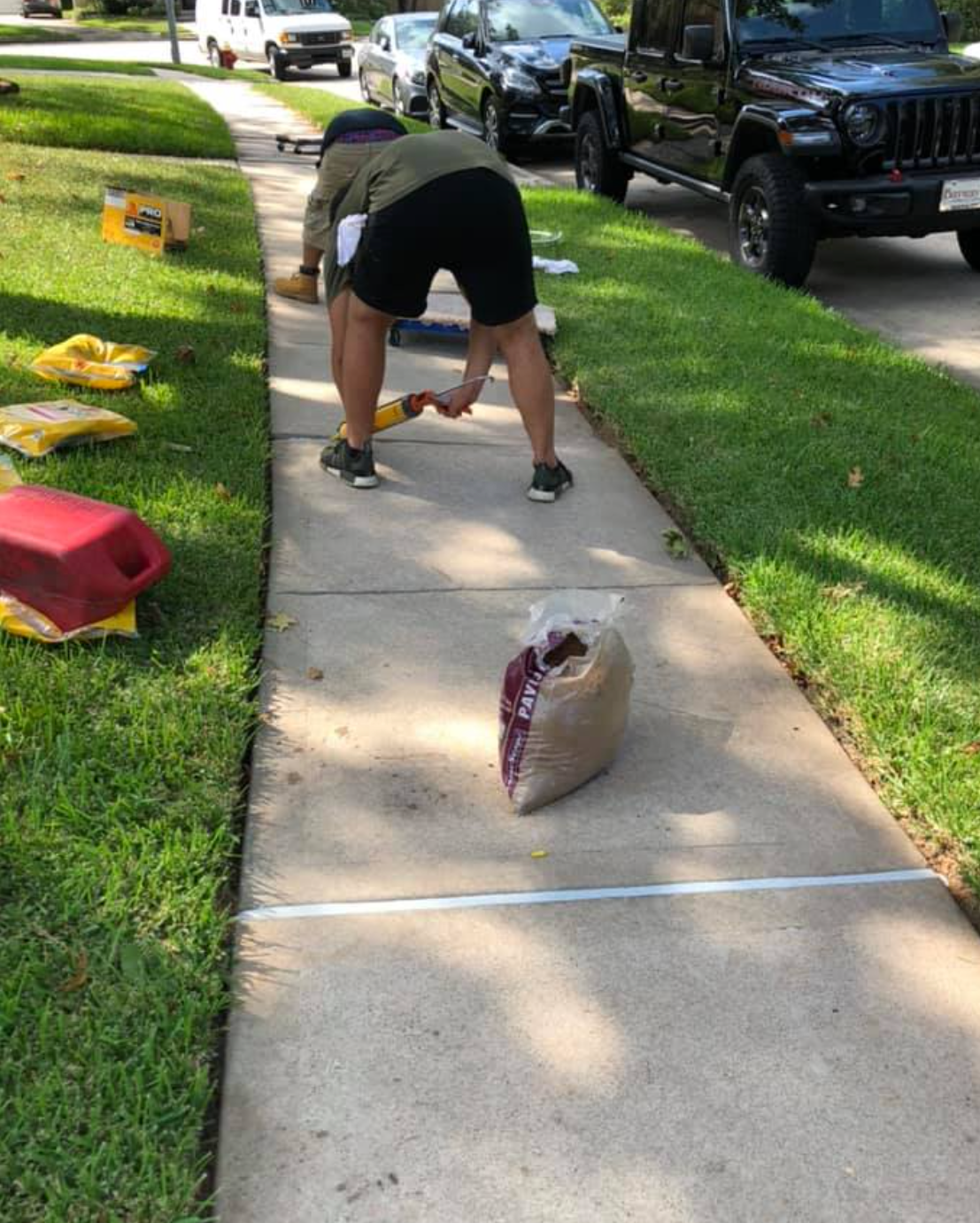 Person repairing a sidewalk, using tools to add material to a marked section; supplies and vehicles are nearby.