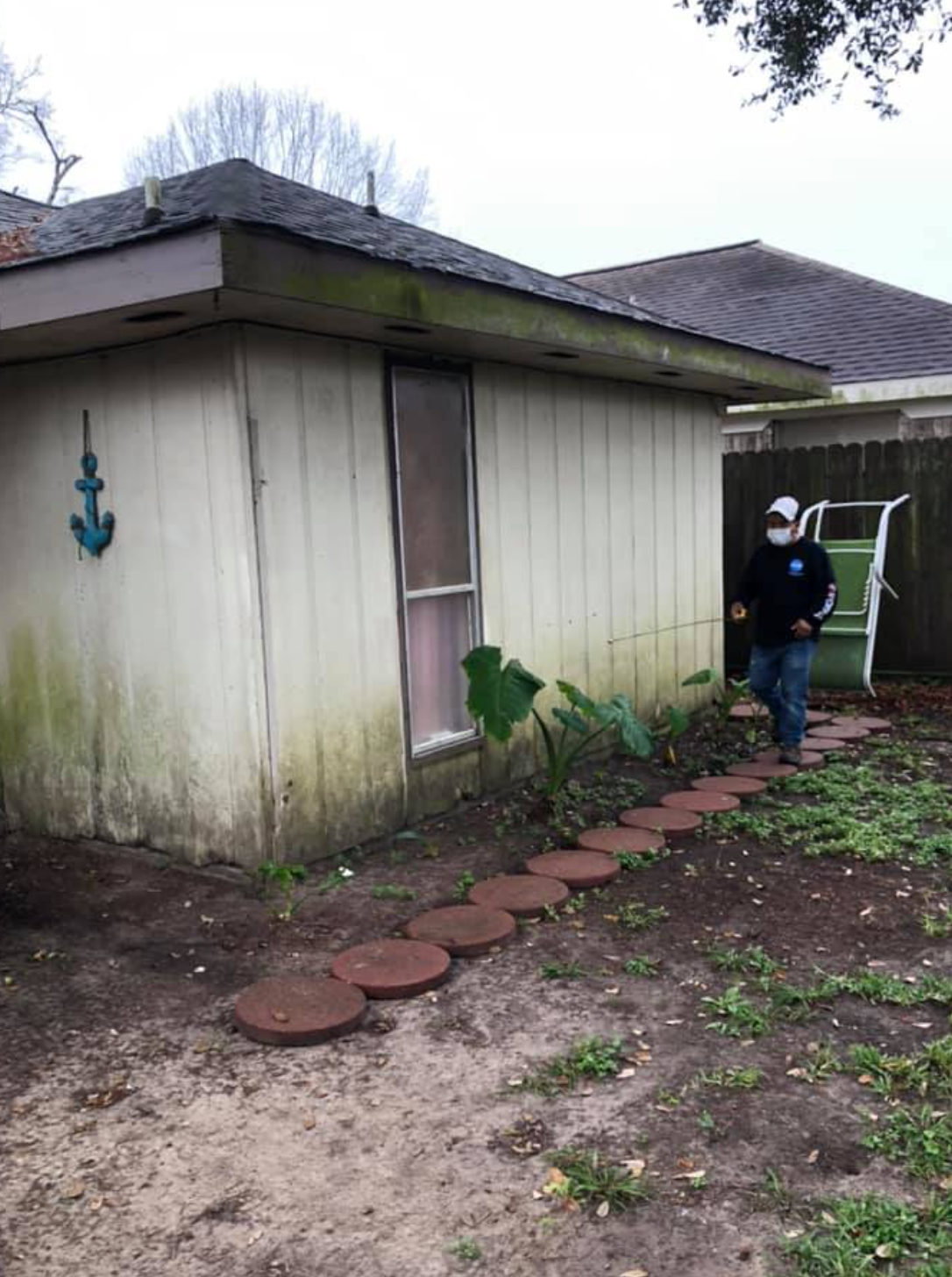 Person walks a brick path beside a weathered, beige building with a window; an anchor decoration hangs outside.