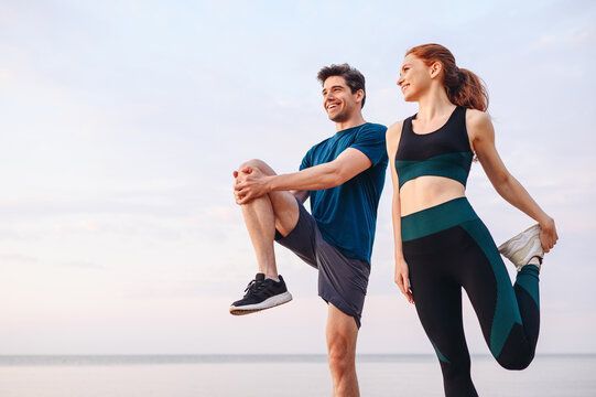 Man and woman stretching legs outdoors, smiling. Cloudy sky in the background.