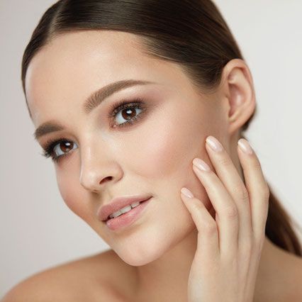 Woman with glowing skin and makeup, touching her face, light background.