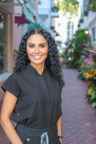 Woman with curly dark hair smiles in a brick alleyway, wearing black scrubs.