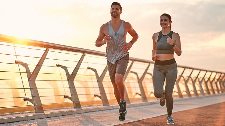 Man and woman running together on a bridge, smiling at sunrise.