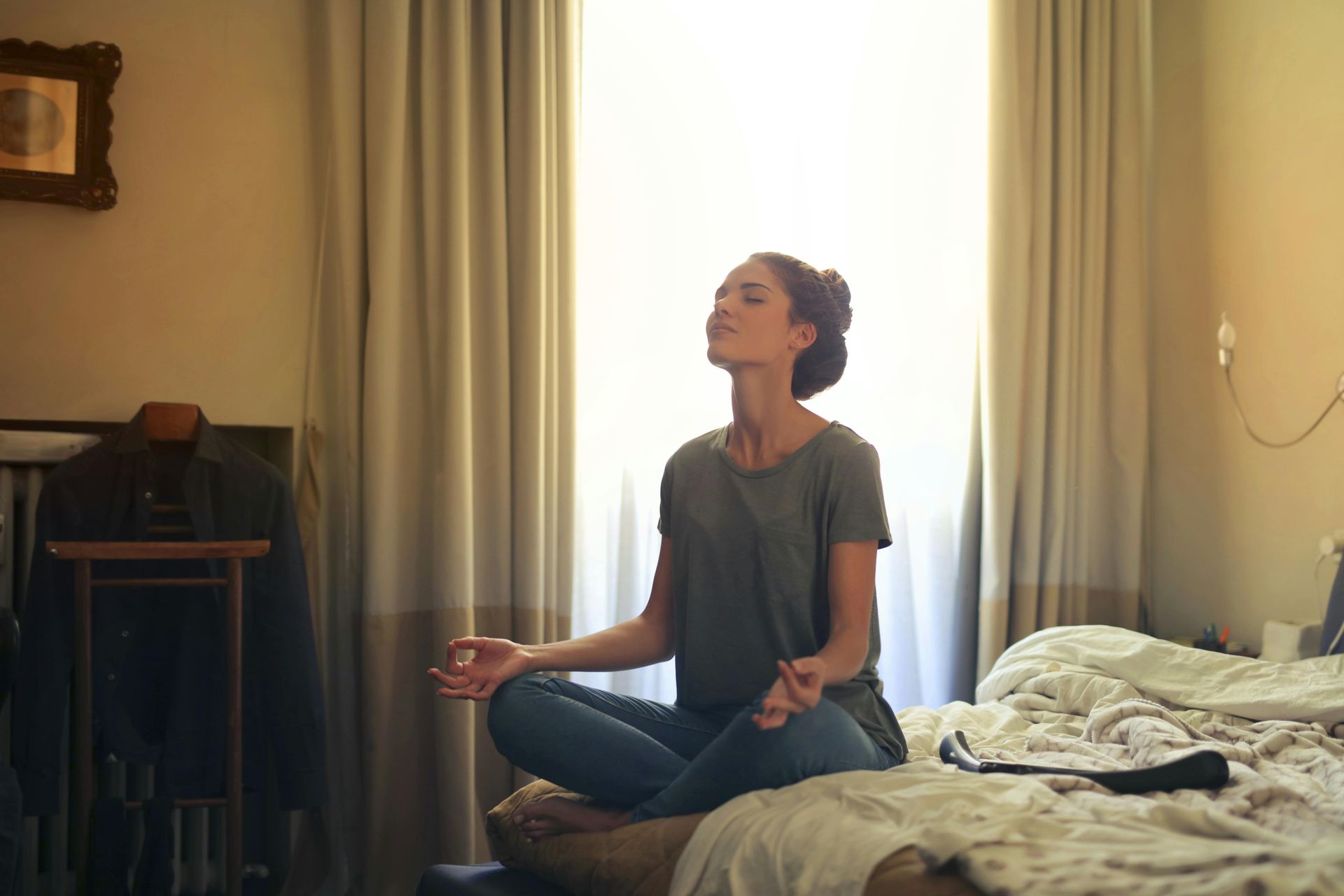 Woman meditating with eyes closed, sitting cross-legged on bed in a room with light from a window.