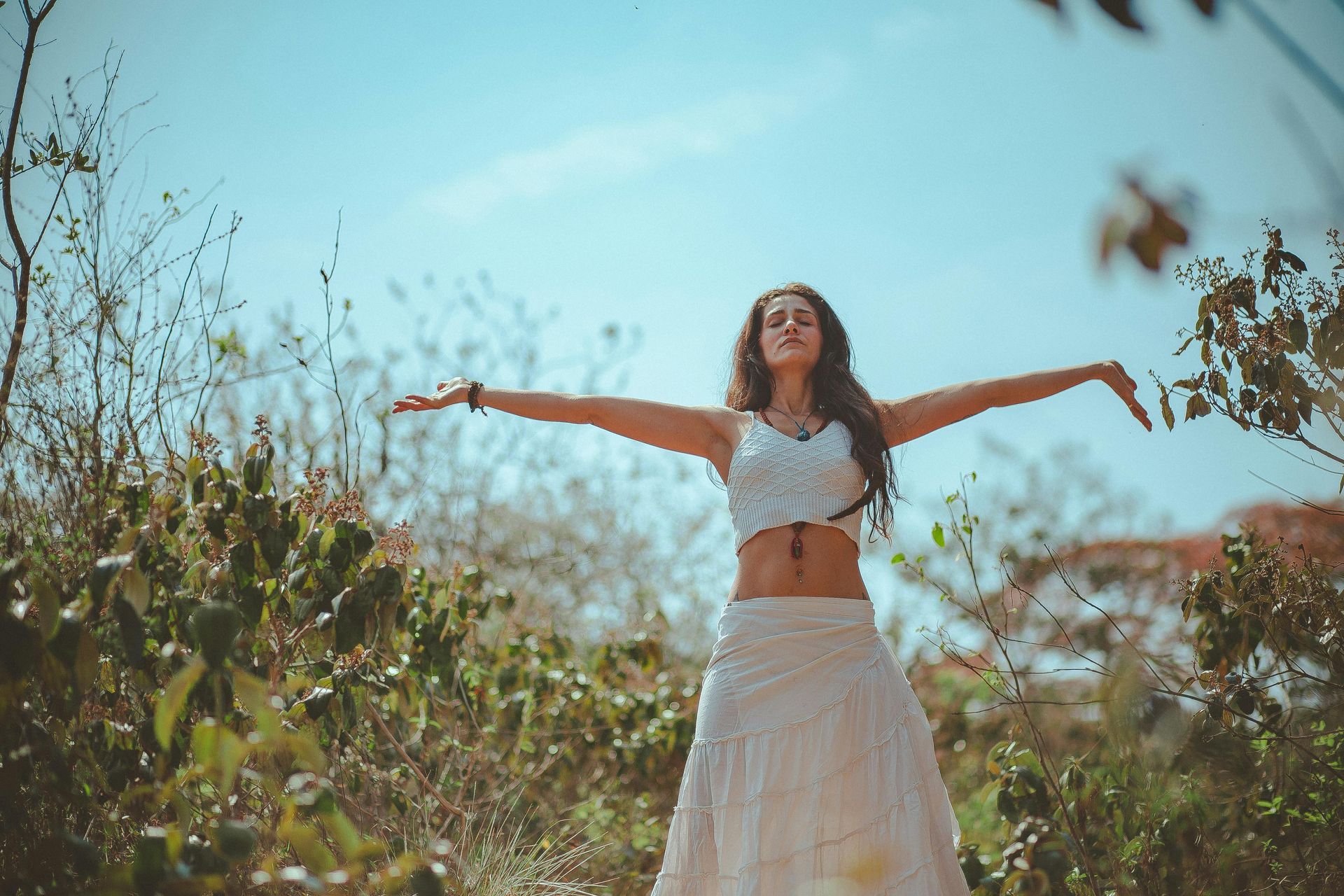 Woman with arms outstretched in a field, wearing white crop top and skirt, blue sky overhead.