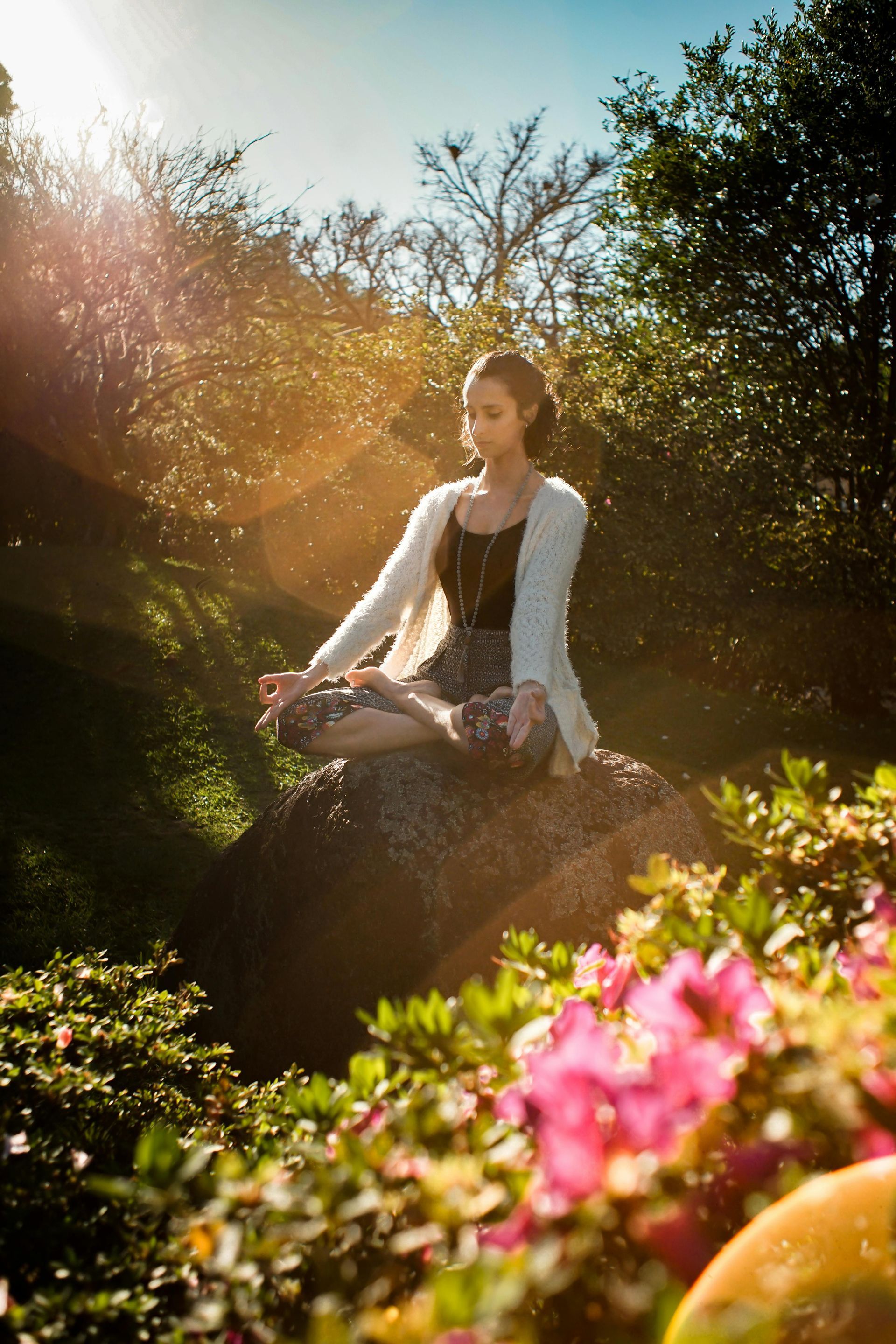 Woman meditating cross-legged on a rock in a sunny garden, surrounded by flowers.
