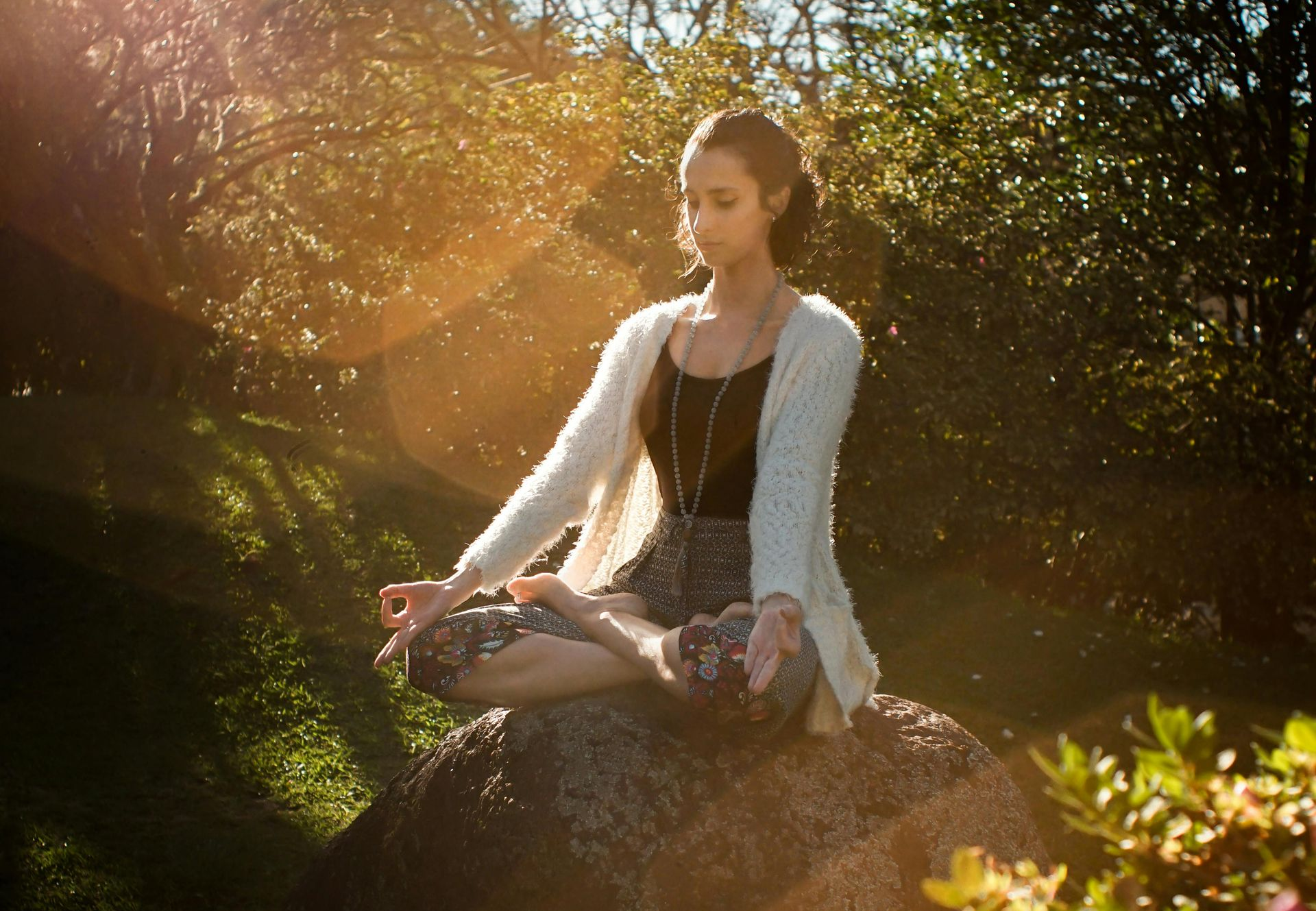 Woman meditating outdoors in a garden, sunlight streaming through trees. She sits cross-legged on a rock.