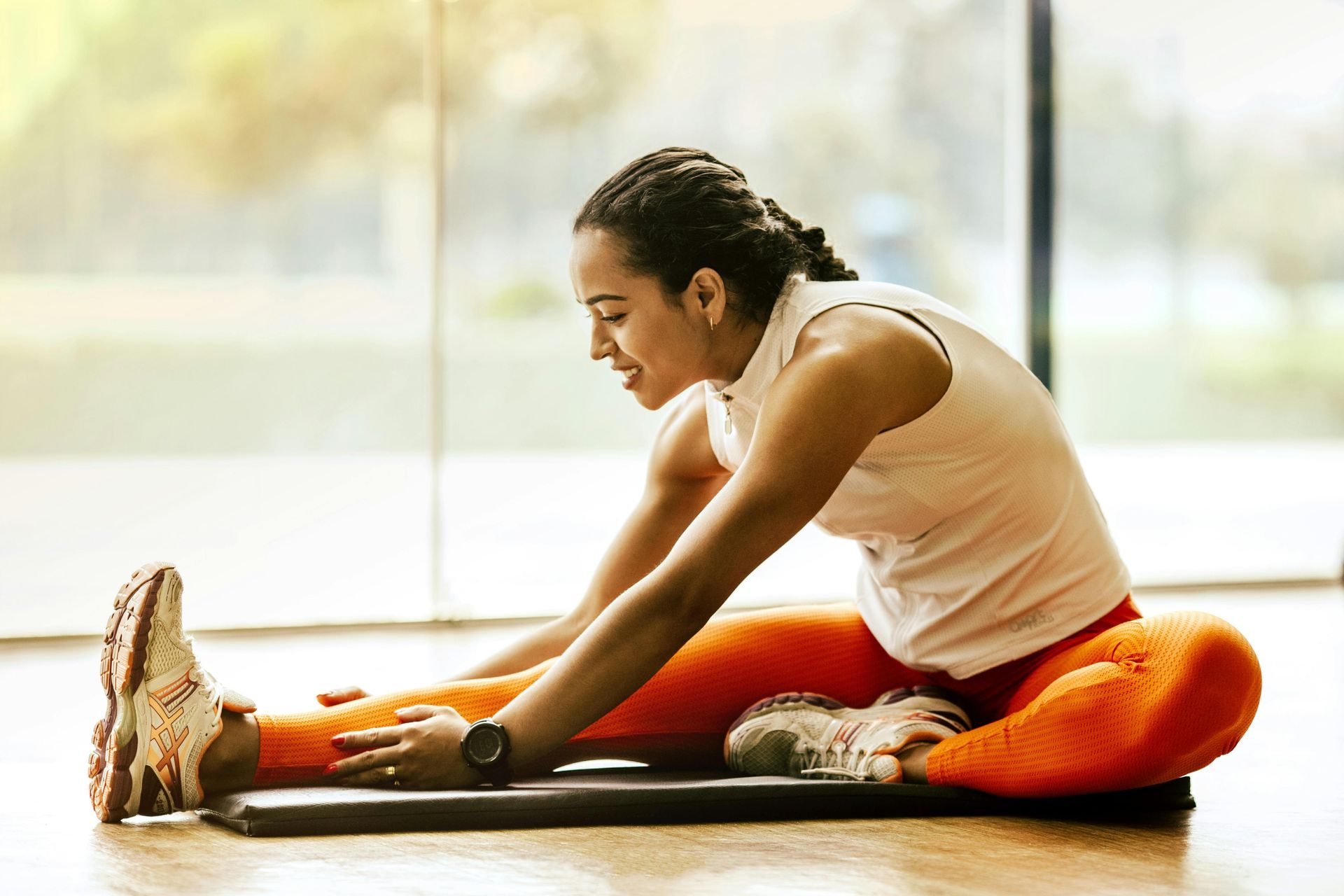 Woman stretching legs while sitting on exercise mat; wearing orange pants and white top.
