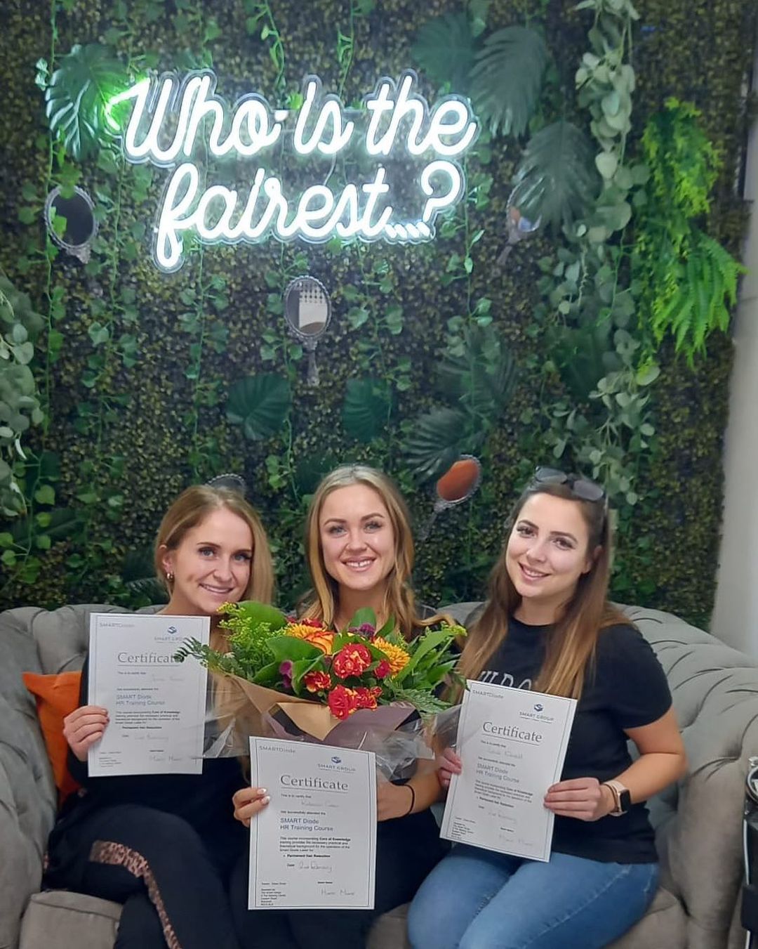 Three women are sitting on a couch holding flowers and certificates.