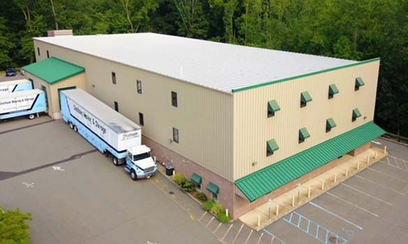 A semi-truck loading at a light tan warehouse with green trim and a paved parking lot.
