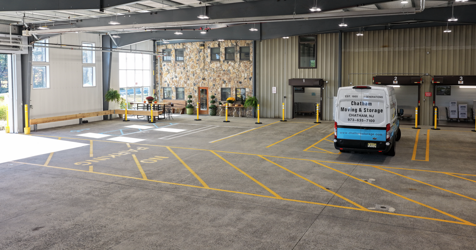 A semi-truck loading at a light tan warehouse with green trim and a paved parking lot.
