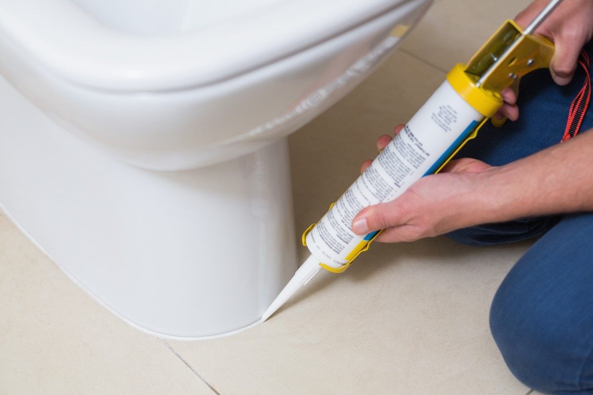 Person applying sealant to the base of a white toilet.