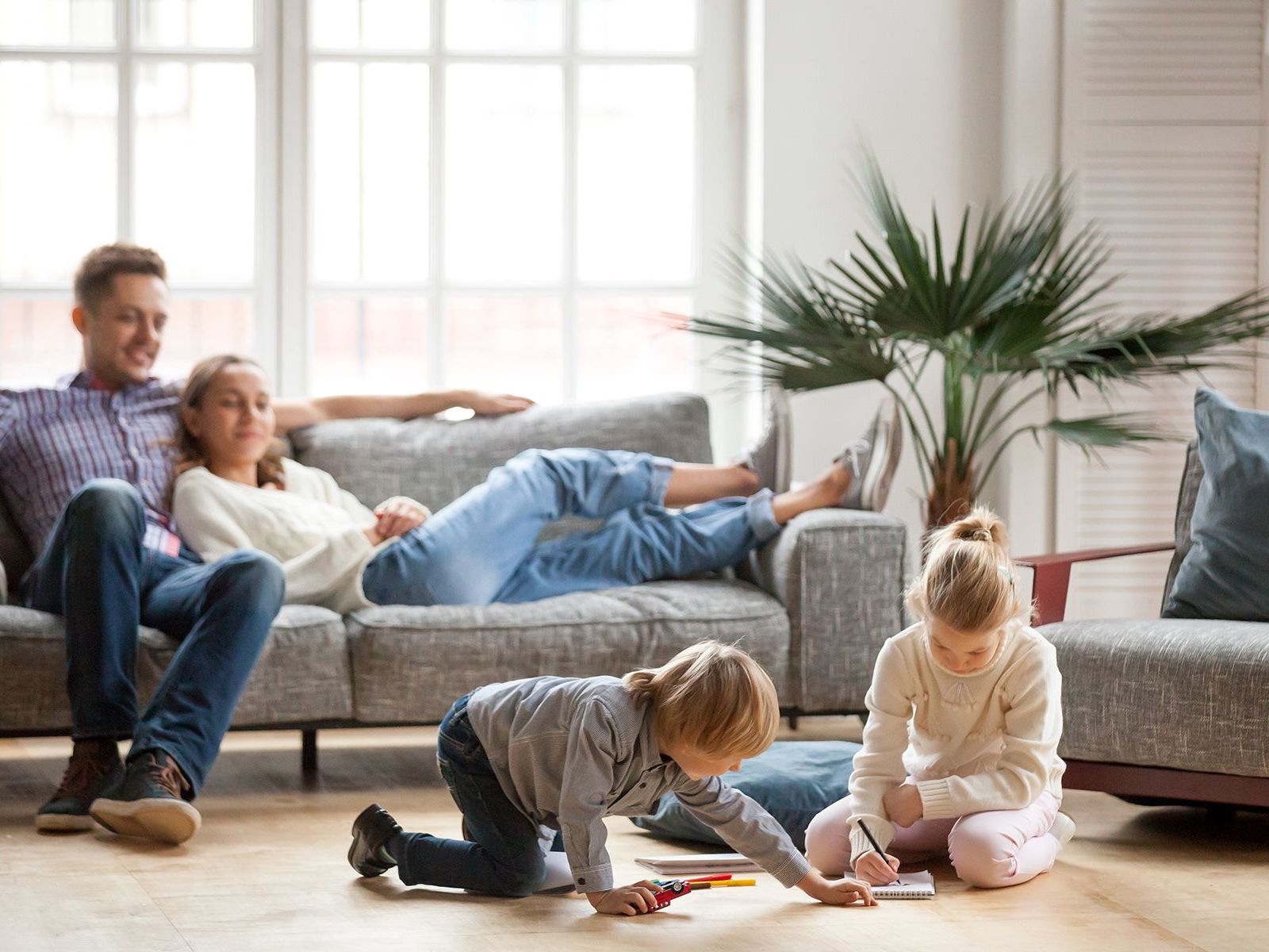 Family relaxing in living room: parents on couch, children playing on floor.