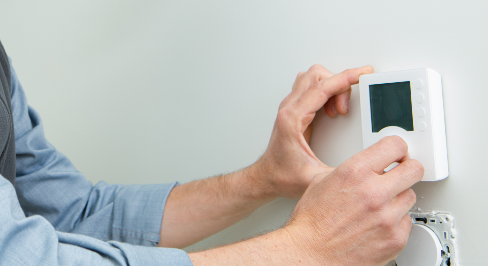 Person adjusting a white thermostat on a white wall, with a light blue shirt sleeve visible.
