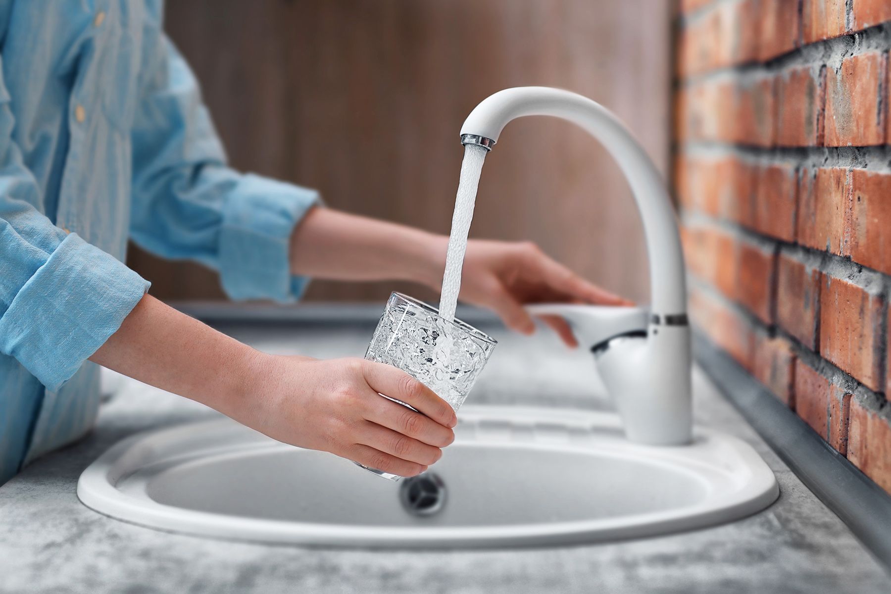 Person filling a glass with water from a white faucet over a white sink. Brick wall on the right.