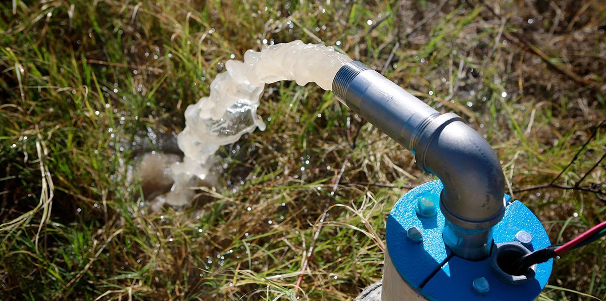 A well pump expels water onto grassy ground, blue base and grey pipe.