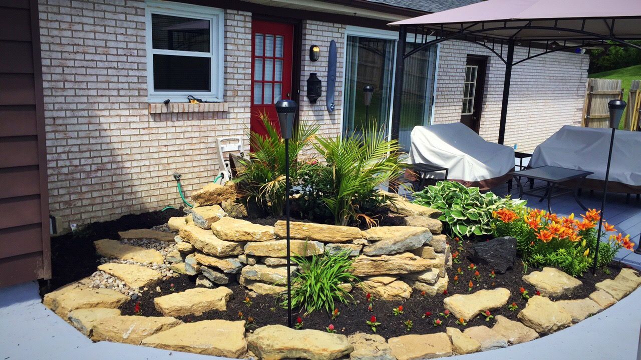 Backyard with a stone-edged flower bed, featuring ferns and lilies, against a brick building with a red door.