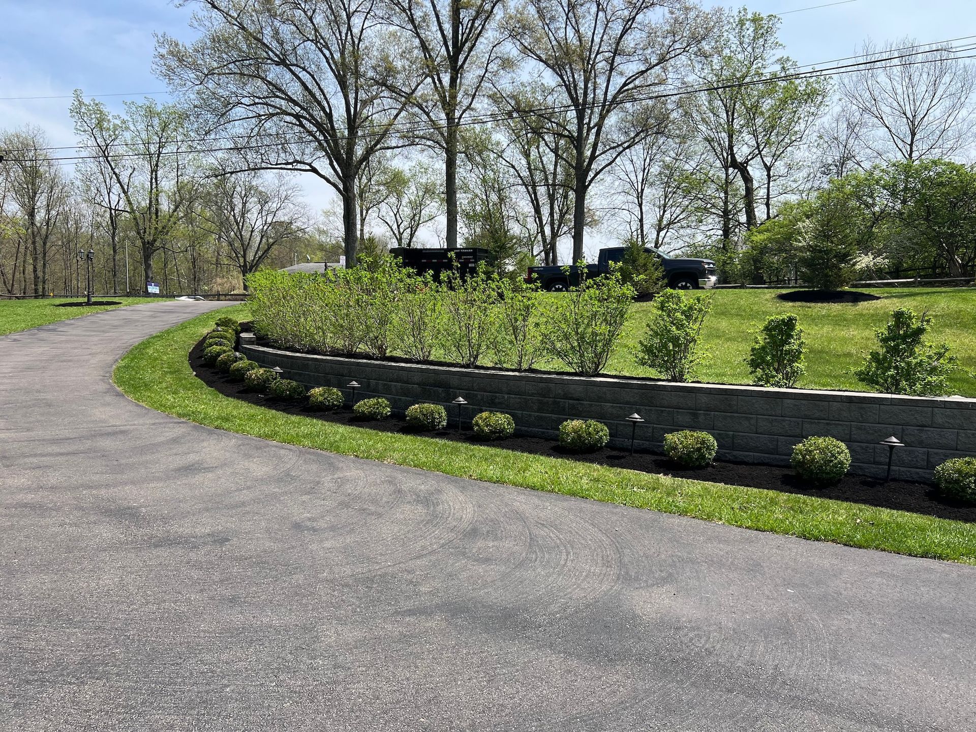 Curved driveway next to a brick retaining wall with bushes and small green shrubs, trees in the background.