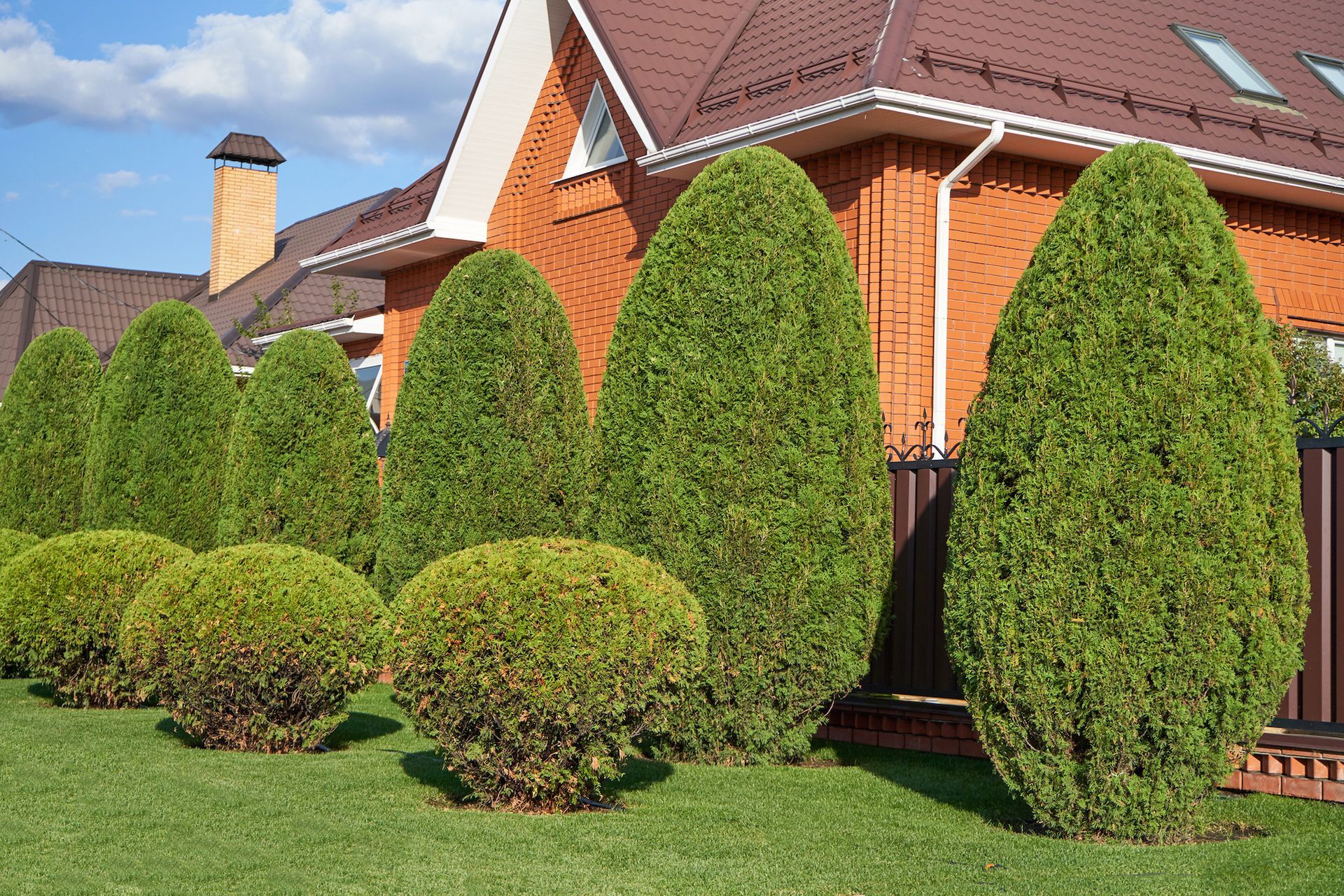 Lush green manicured bushes and trees border a red brick house with a brown roof and brown fence.