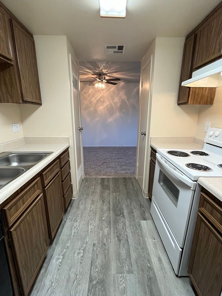 Kitchen with brown cabinets, white appliances, and a doorway to a living area with gray carpet.
