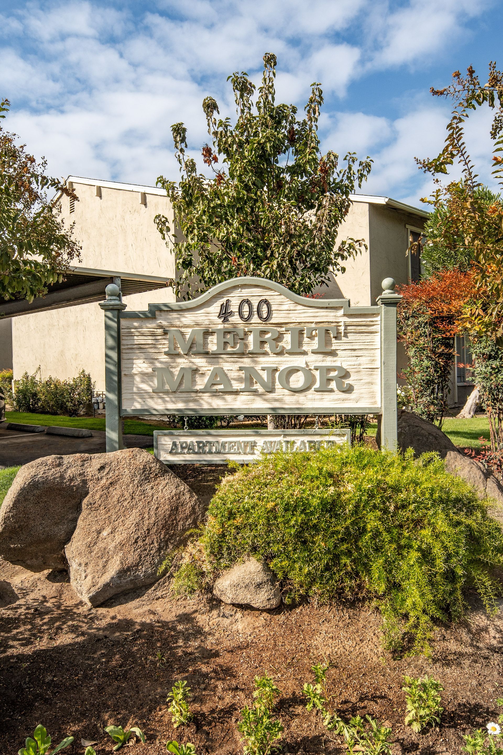 Sign for Merit Manor apartments with landscaping; light brown sign with green lettering, set in front of tan buildings.