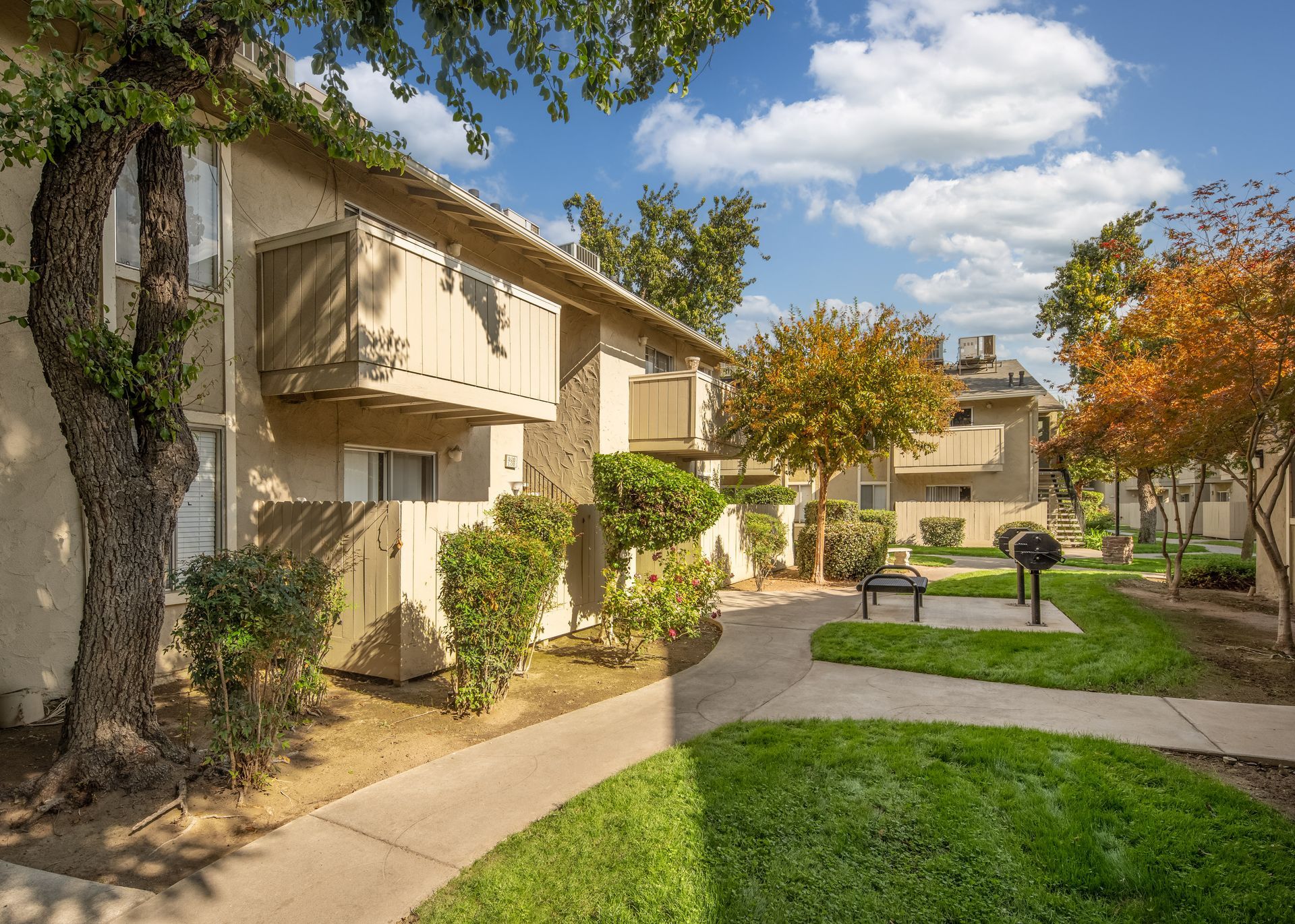 Apartment complex with beige buildings, green lawn, and walkway. Blue sky and trees.