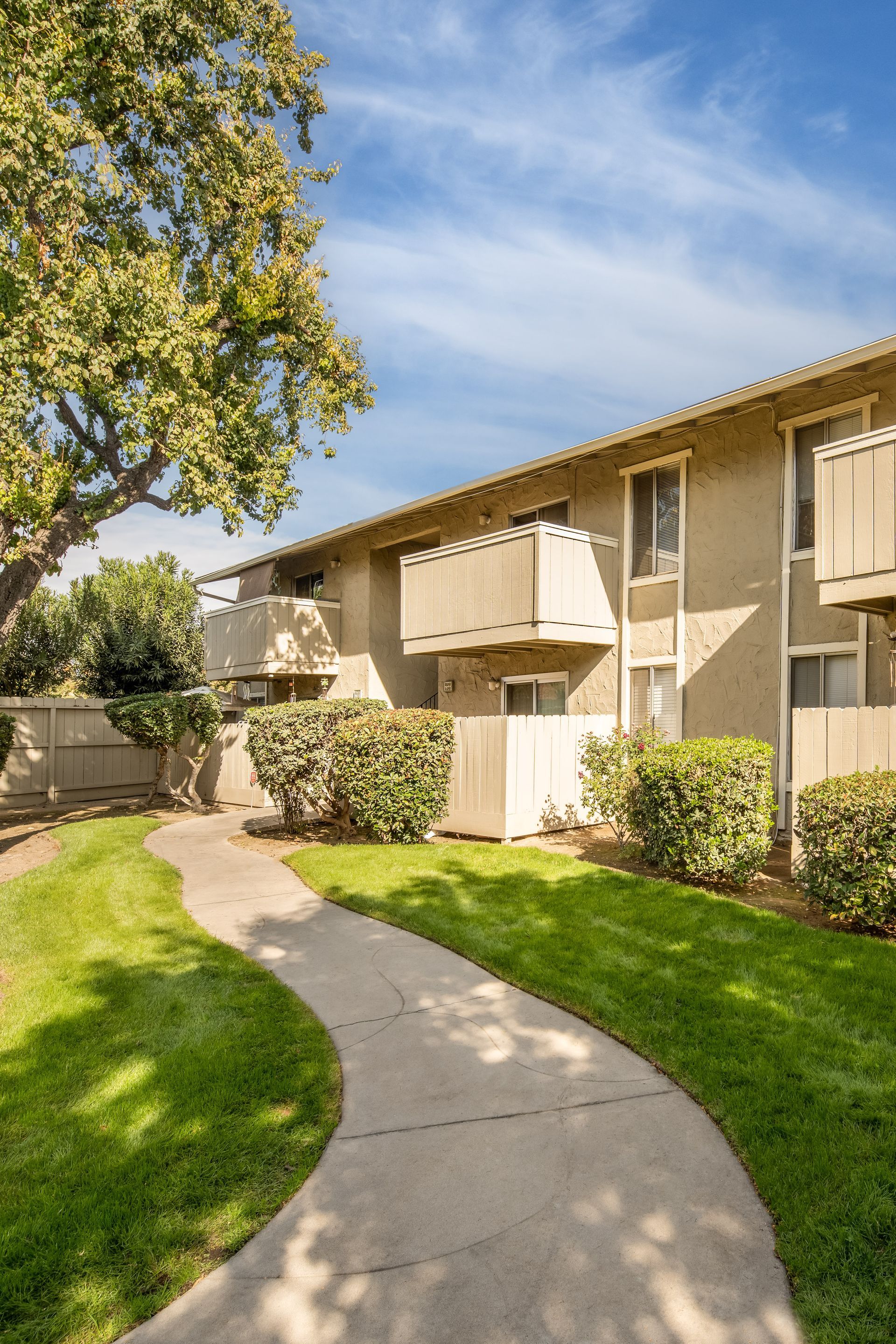 Concrete path winds through green lawn past a two-story apartment building with balconies, under a blue sky.