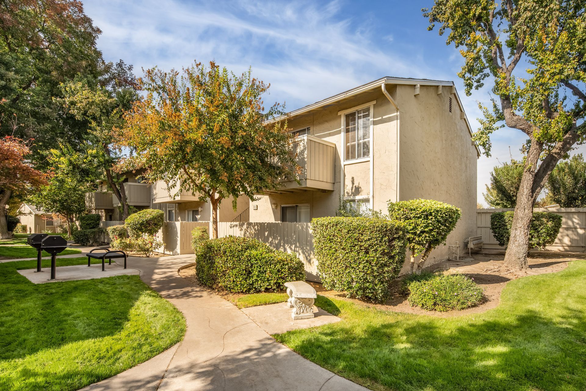 Two-story tan building with trees, grass, pathway, benches, and barbecue on a sunny day.