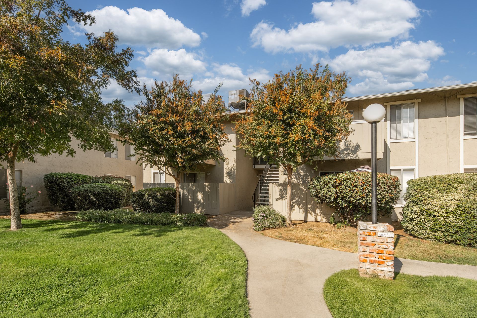 Apartment building with a grassy lawn, trees, and a pathway on a sunny day.