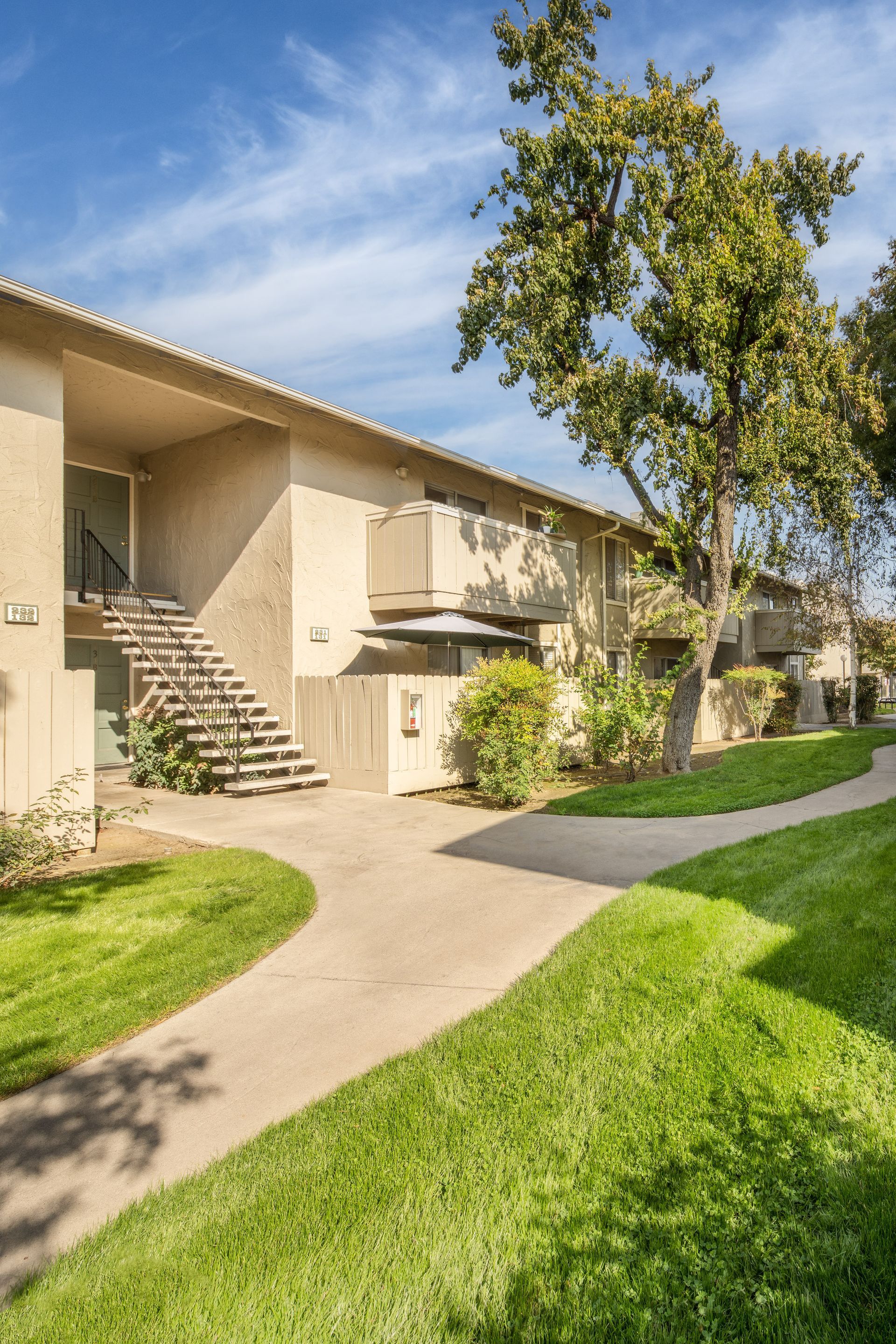 Apartment building exterior with stairs, balconies, and green grass pathway under blue sky.
