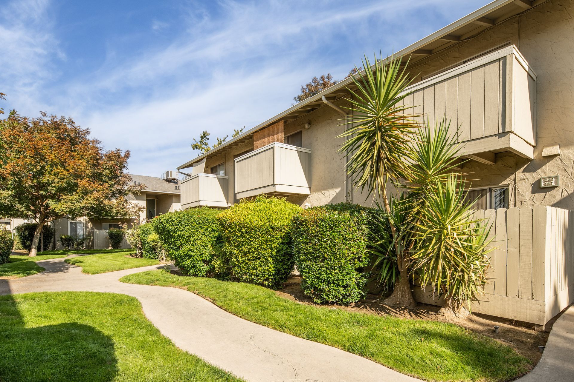 Apartment complex exterior with pathway, green grass, shrubs, and beige buildings. Sunny day.
