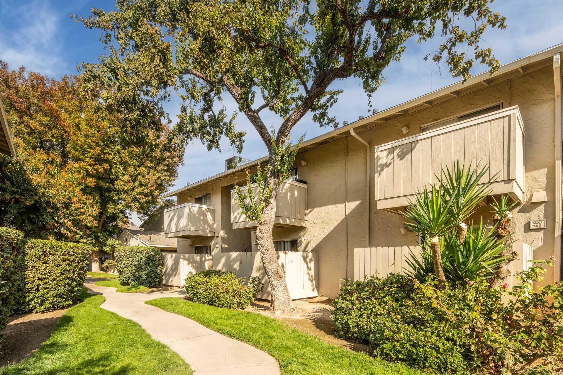 Apartment complex with tan siding, balconies, and pathway lined with greenery.