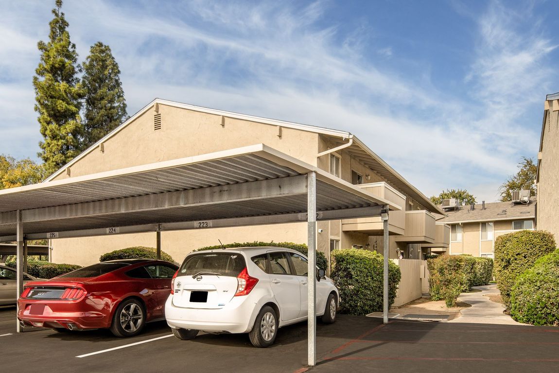 Cars parked under a metal carport outside a two-story apartment building on a sunny day.