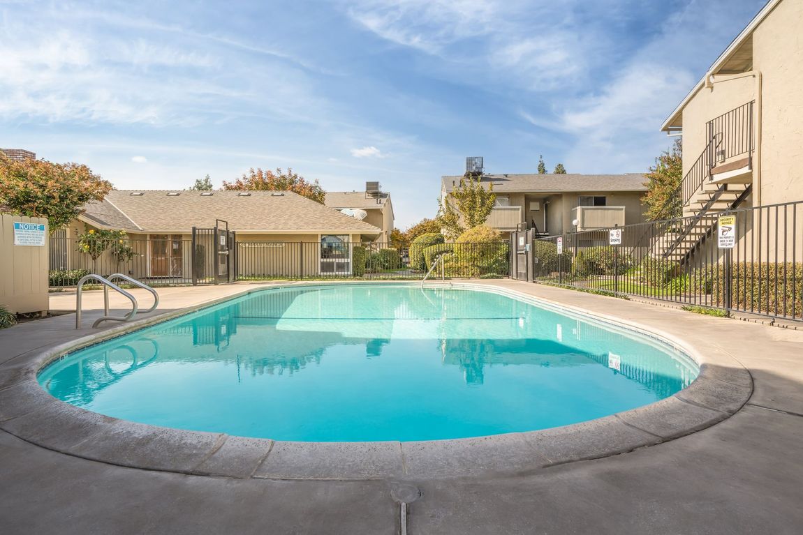 Swimming pool in an apartment complex with blue water, surrounded by buildings and a fence, under a blue sky.