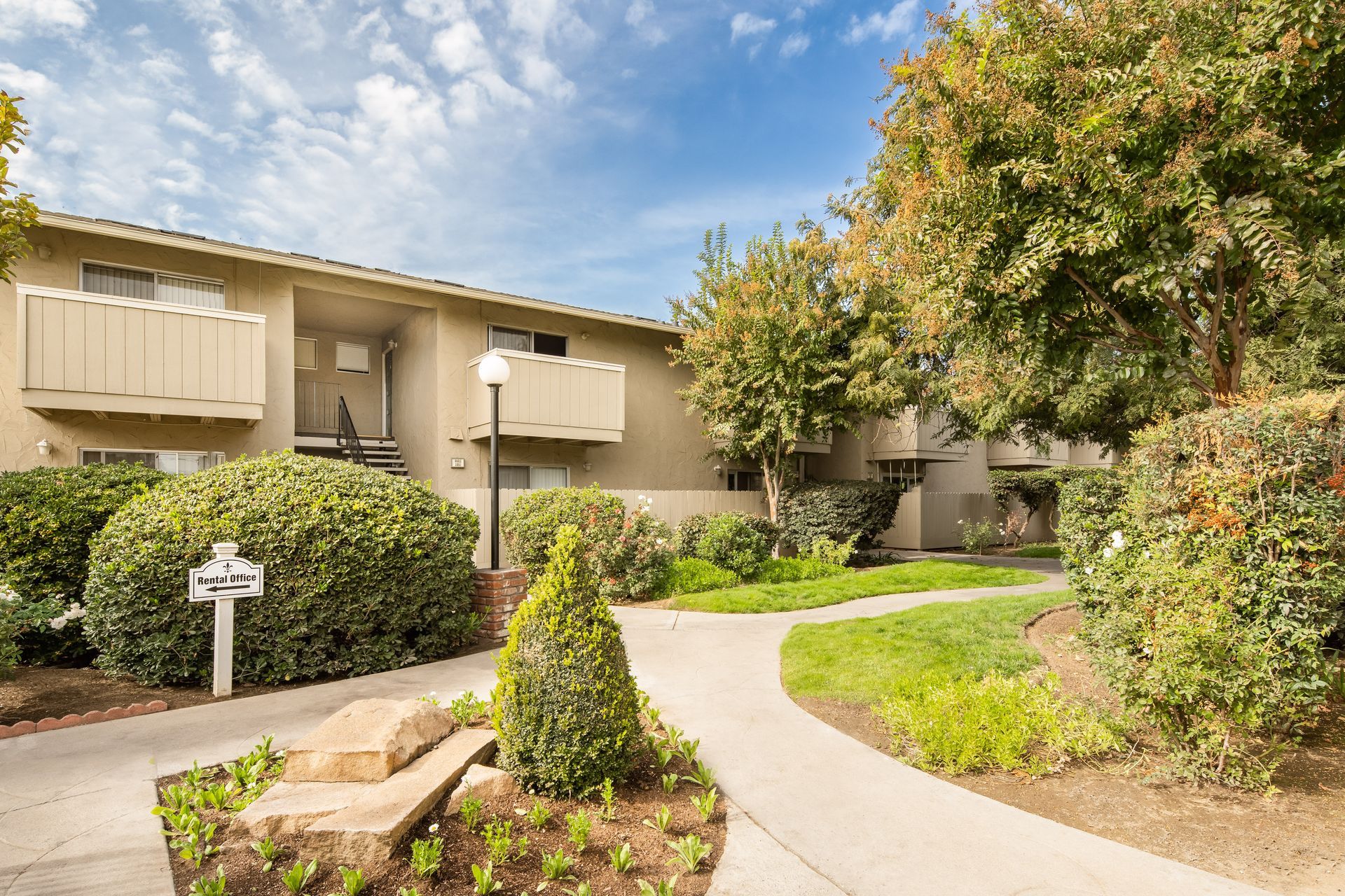 Apartment building exterior with beige facade, balconies, pathway, and lush landscaping under a cloudy sky.