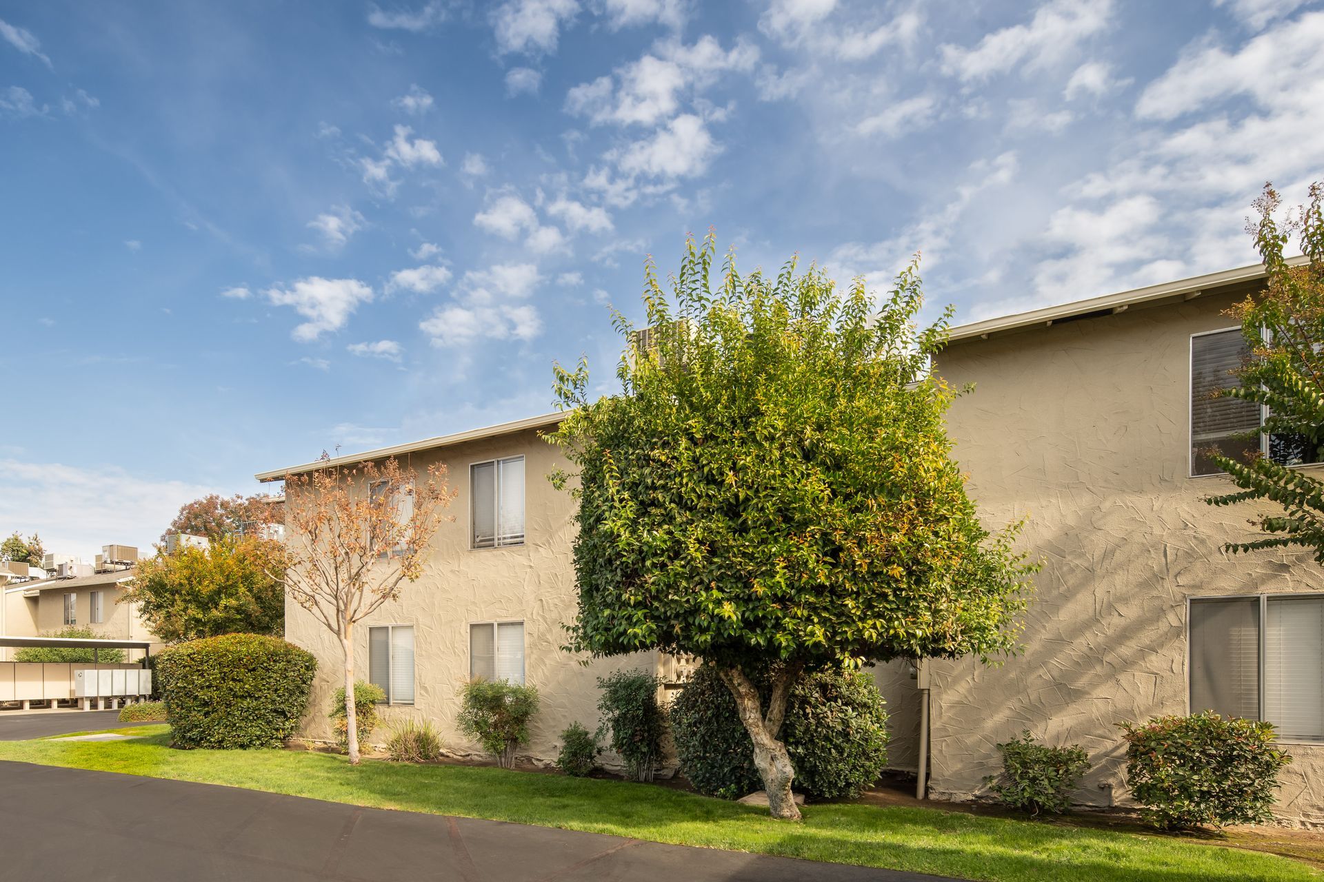 Apartment building with tan stucco exterior, green trees, and blue sky with clouds.
