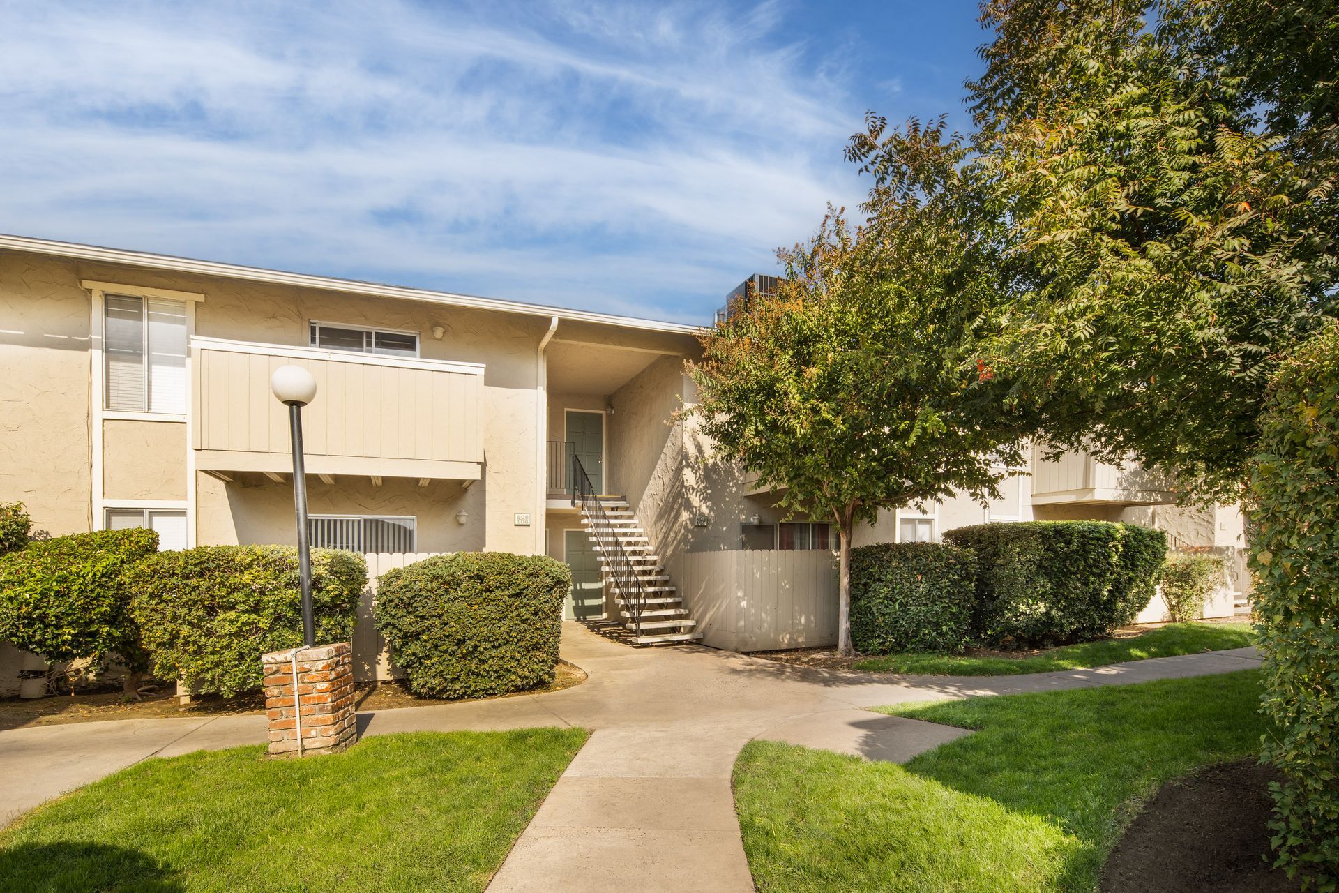 Apartment building exterior with stairs, landscaping, and a walkway under a blue sky.