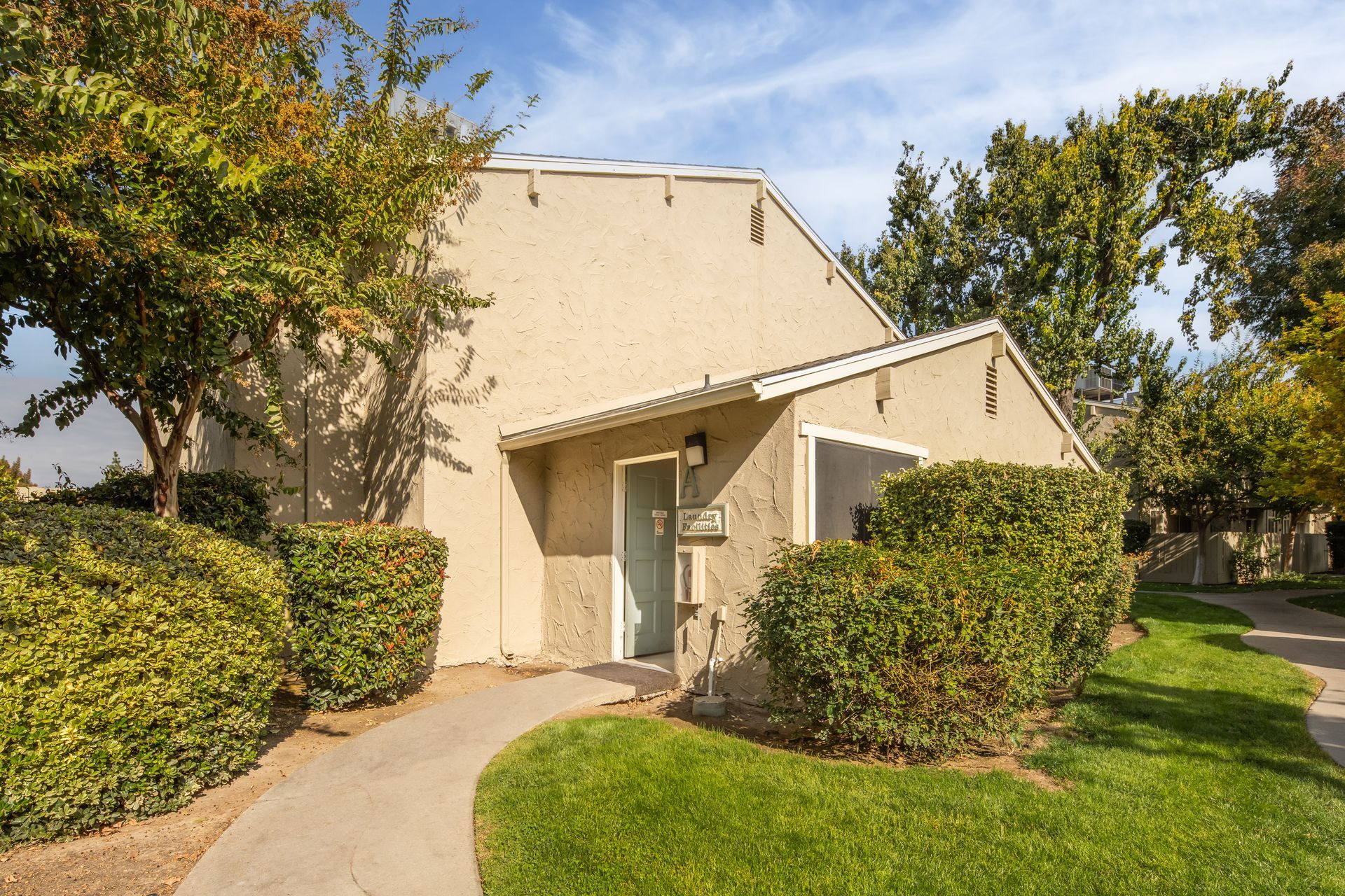 Tan building with a blue door, surrounded by green bushes and grass, a walkway leads to the entrance.