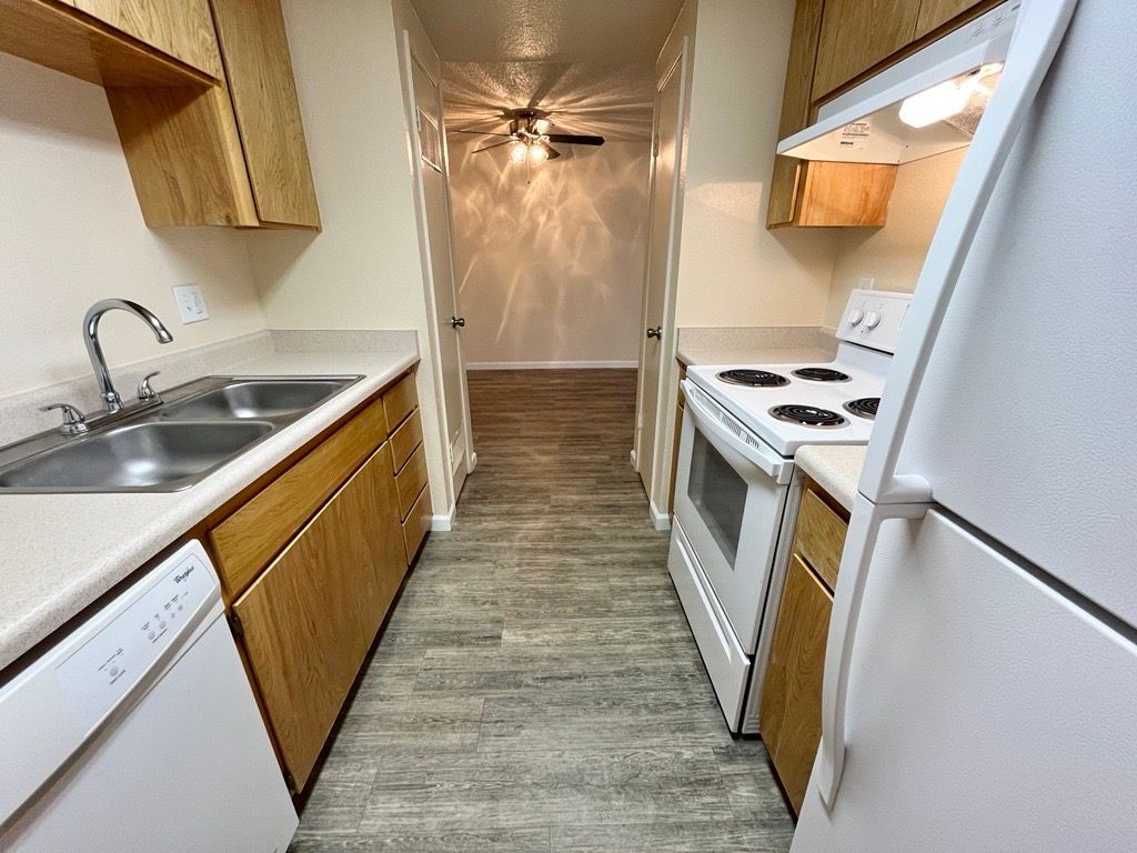 A narrow kitchen with light wood cabinets, white appliances, and a view into a room with a ceiling fan.