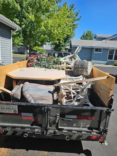 A black trailer is filled with old appliances like a washing machine and a refrigerator. The trailer is parked on a sunny street.