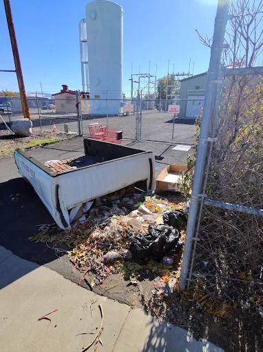 Black utility trailer with a tarp, parked on a paved road with a curb. The trailer has wooden sideboards.