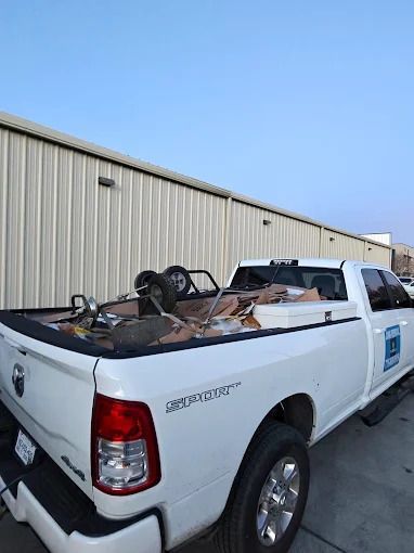 White pickup truck with a bed full of debris parked in front of a metal building under a blue sky.