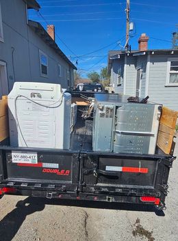 A black trailer is filled with old appliances like a washing machine and a refrigerator. The trailer is parked on a sunny street.