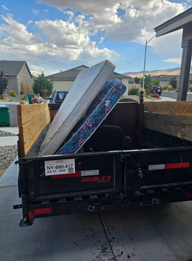 A black trailer filled with discarded mattresses and a chair in a driveway, with houses and blue sky in the background.