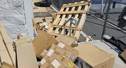 Pile of cardboard boxes and wooden pallets leaning against a brick wall and chain-link fence, outdoors on a sunny day.