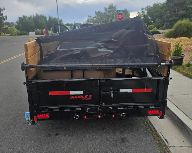 Black utility trailer with a tarp, parked on a paved road with a curb. The trailer has wooden sideboards.