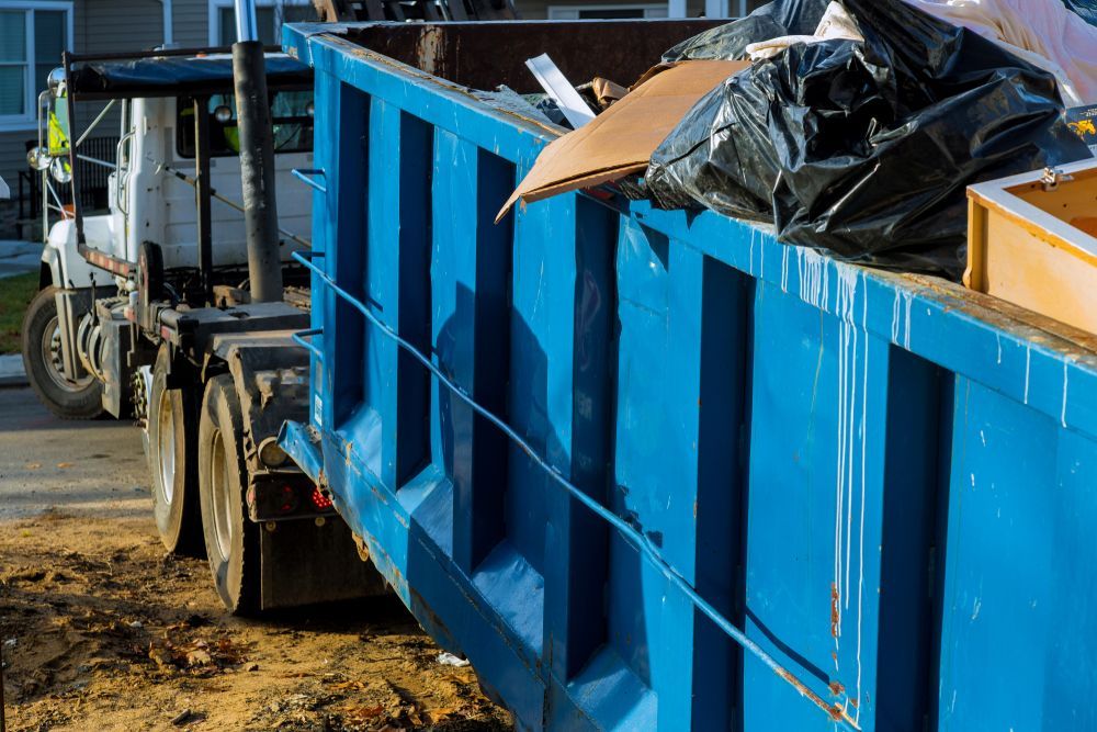 Blue dumpster filled with trash beside a garbage truck, outdoors.