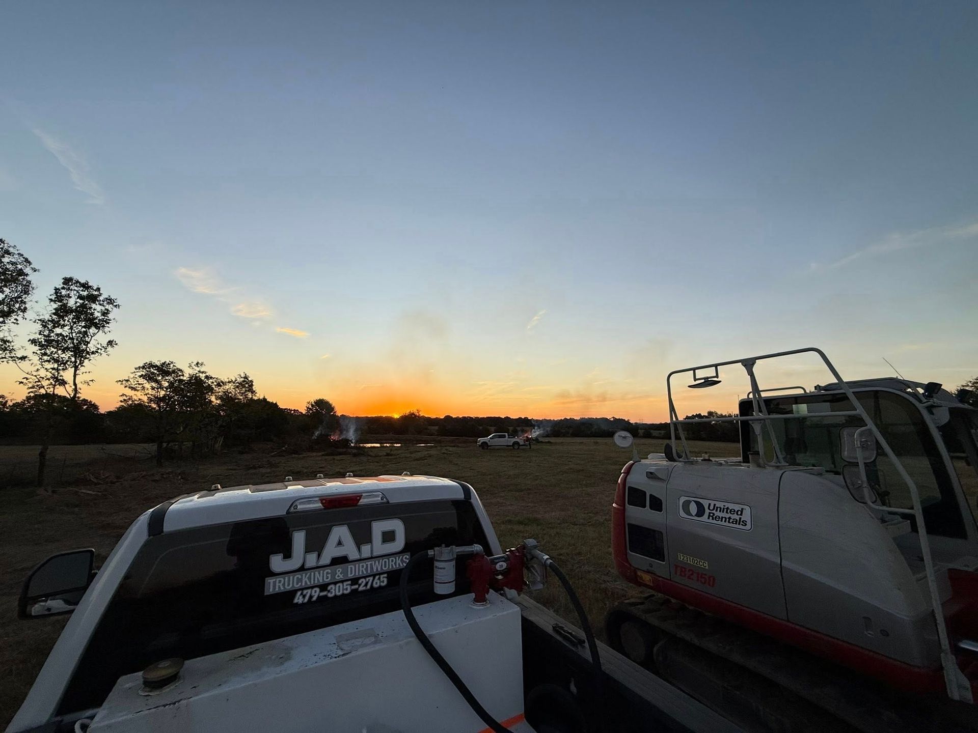 A truck and excavator sit in a field at sunset; golden sky, trees, and distant smoke.