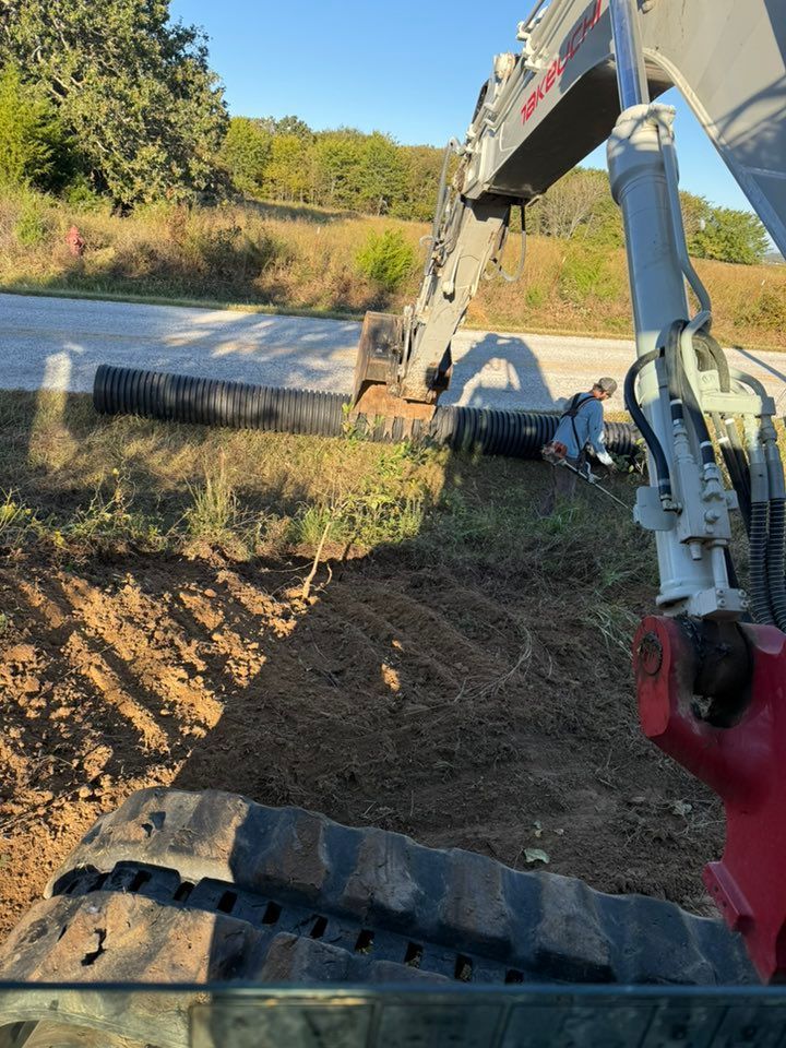 An excavator near black pipes; a person is working in the trench. Sunny day, outdoor setting.